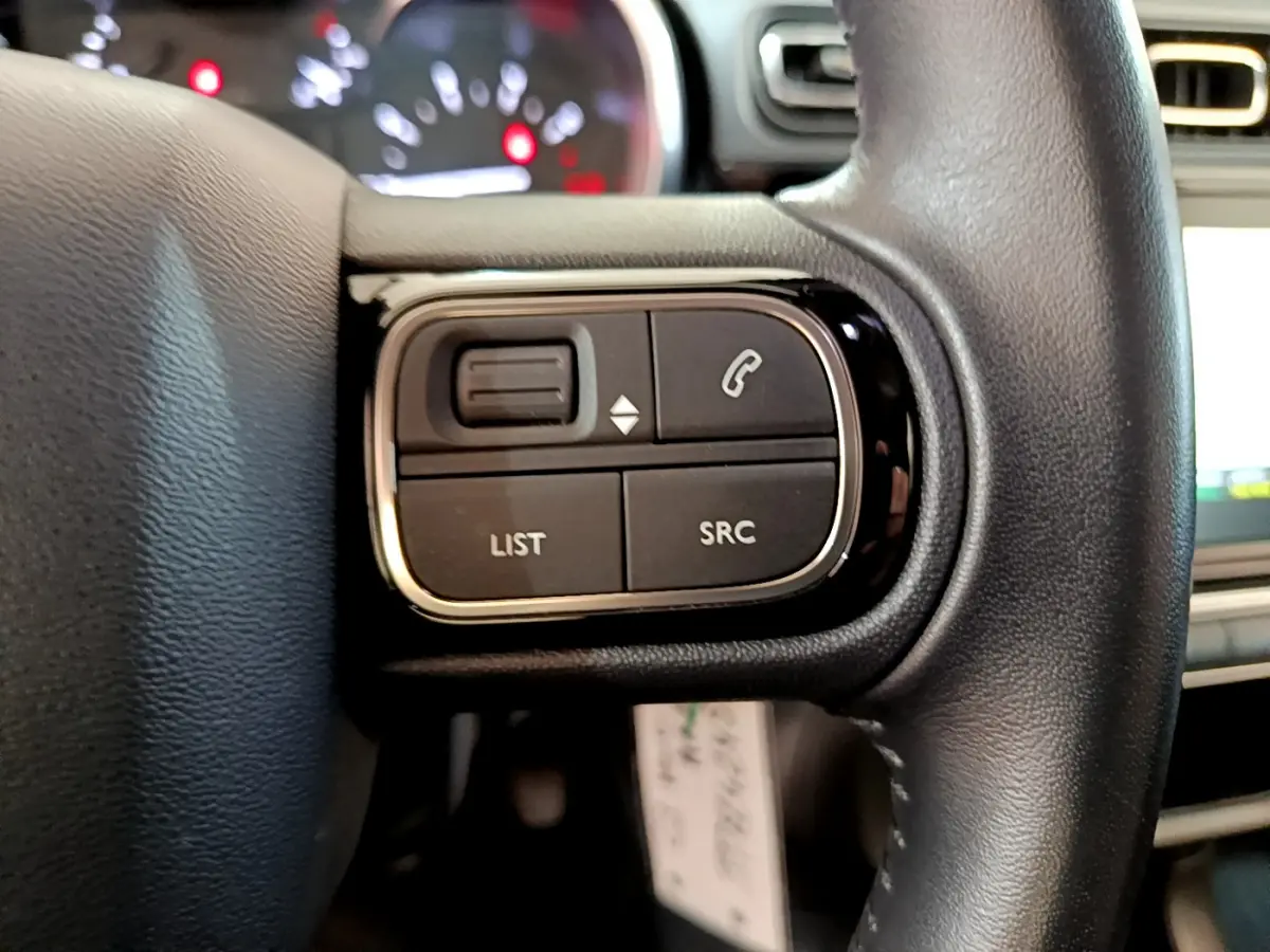 Gros plan sur les commandes du volant cuir côté gauche de la Citroën C3 blanche, avec écran tactile en arrière-plan.