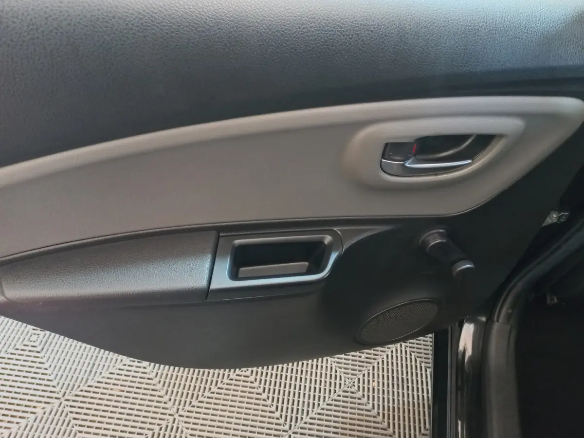 Intérieur côté gauche de la porte d'une Toyota Yaris noire, avec poignée argentée et commande manuelle de vitre.