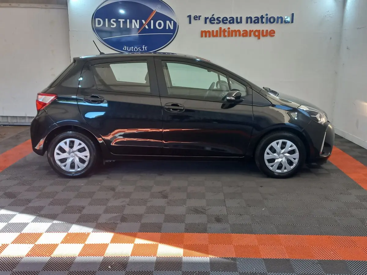 Profil côté gauche d'une Toyota Yaris noire 2018 hybride, stationnée en intérieur sur sol damier gris et orange.