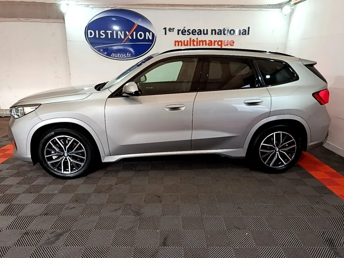 Vue de profil côté gauche d’un BMW X1 xDrive23i M Sport gris clair, roues alliage et vitres teintées, en intérieur showroom.