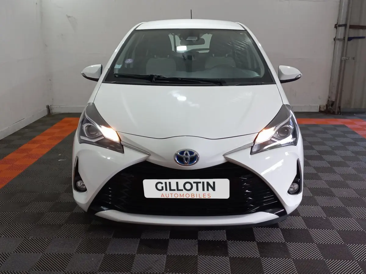 Vue avant d'une Toyota Yaris blanche 2019 avec phares allumés et calandre noire distinctive en intérieur showroom.