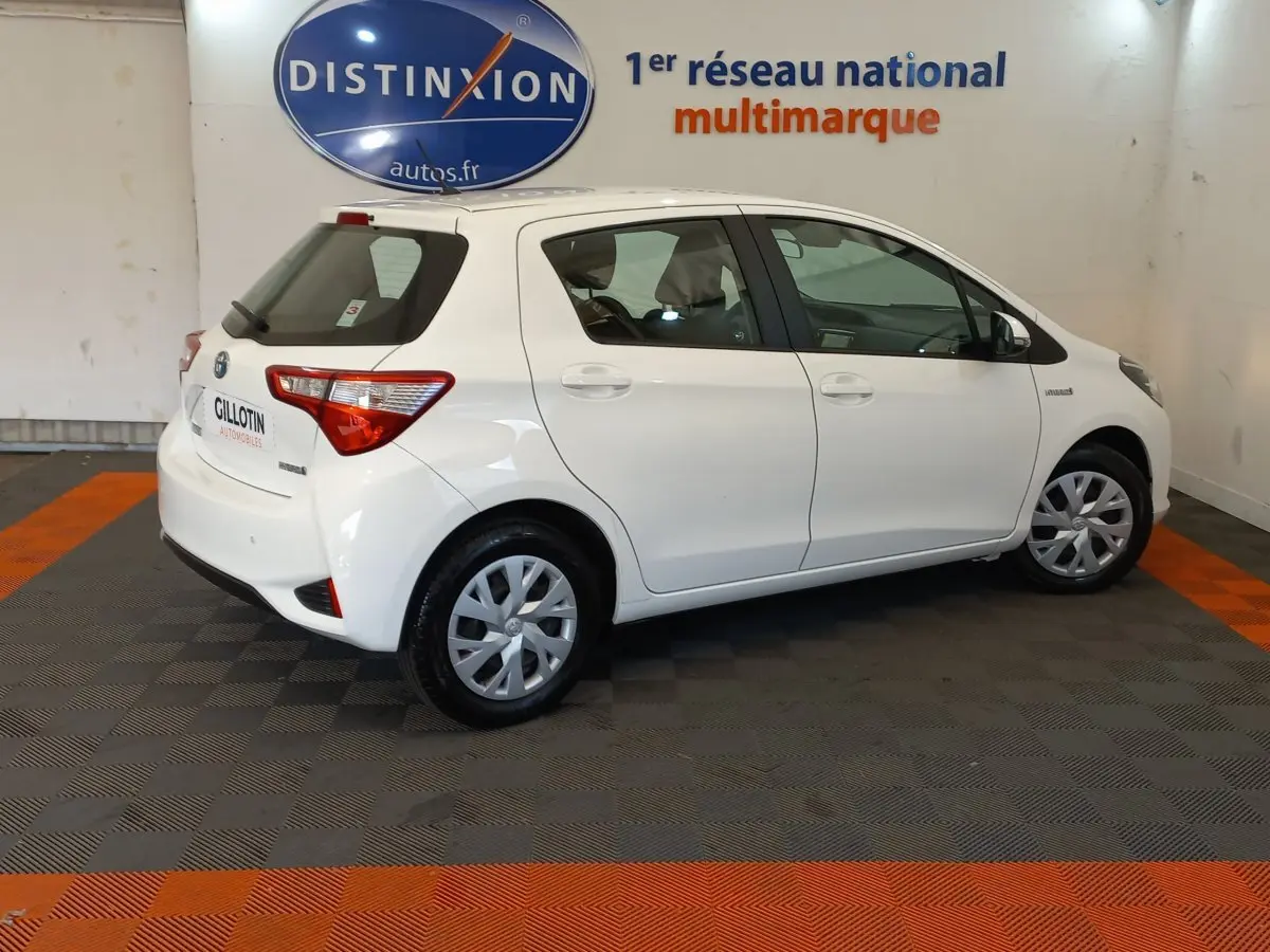 Profil droit d'une Toyota Yaris blanche 2019 hybride, avec jantes standard et logo Distinxion en arrière-plan.