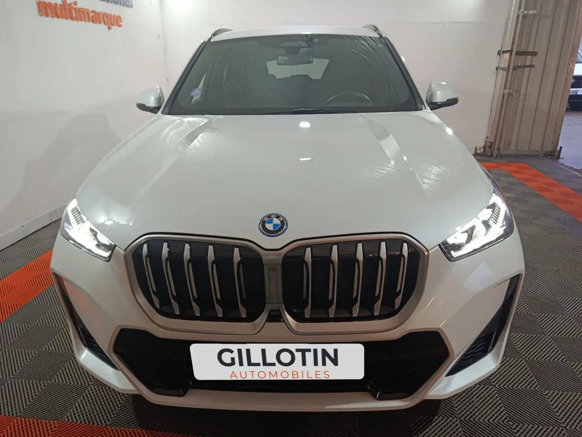 Vue frontale d’un BMW X1 blanc 2024 avec calandre imposante et phares LED allumés dans un garage.