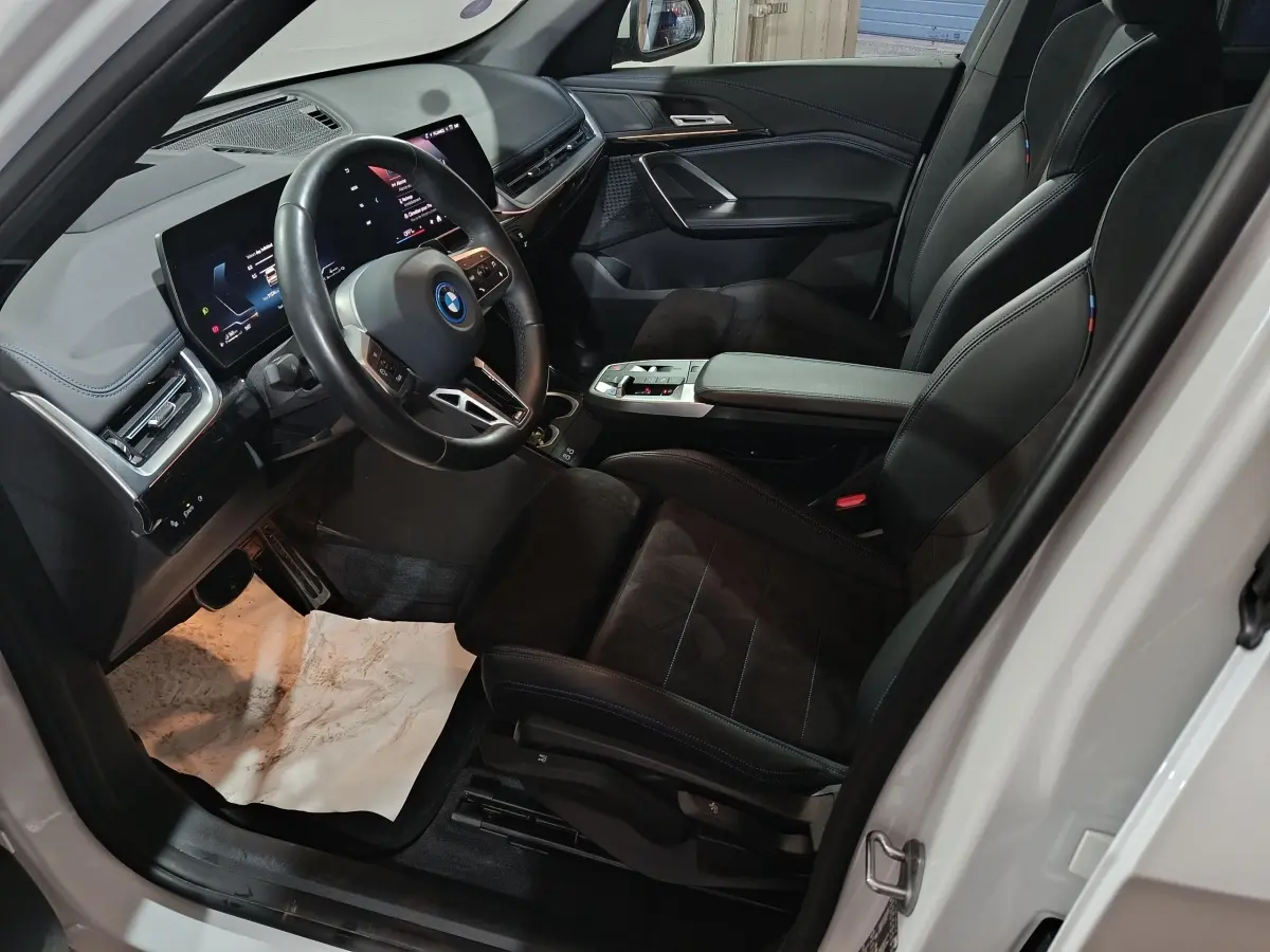 Vue intérieure côté conducteur du BMW X1 xDrive25e blanc 2024, avec tableau de bord digital et sièges sport noirs.