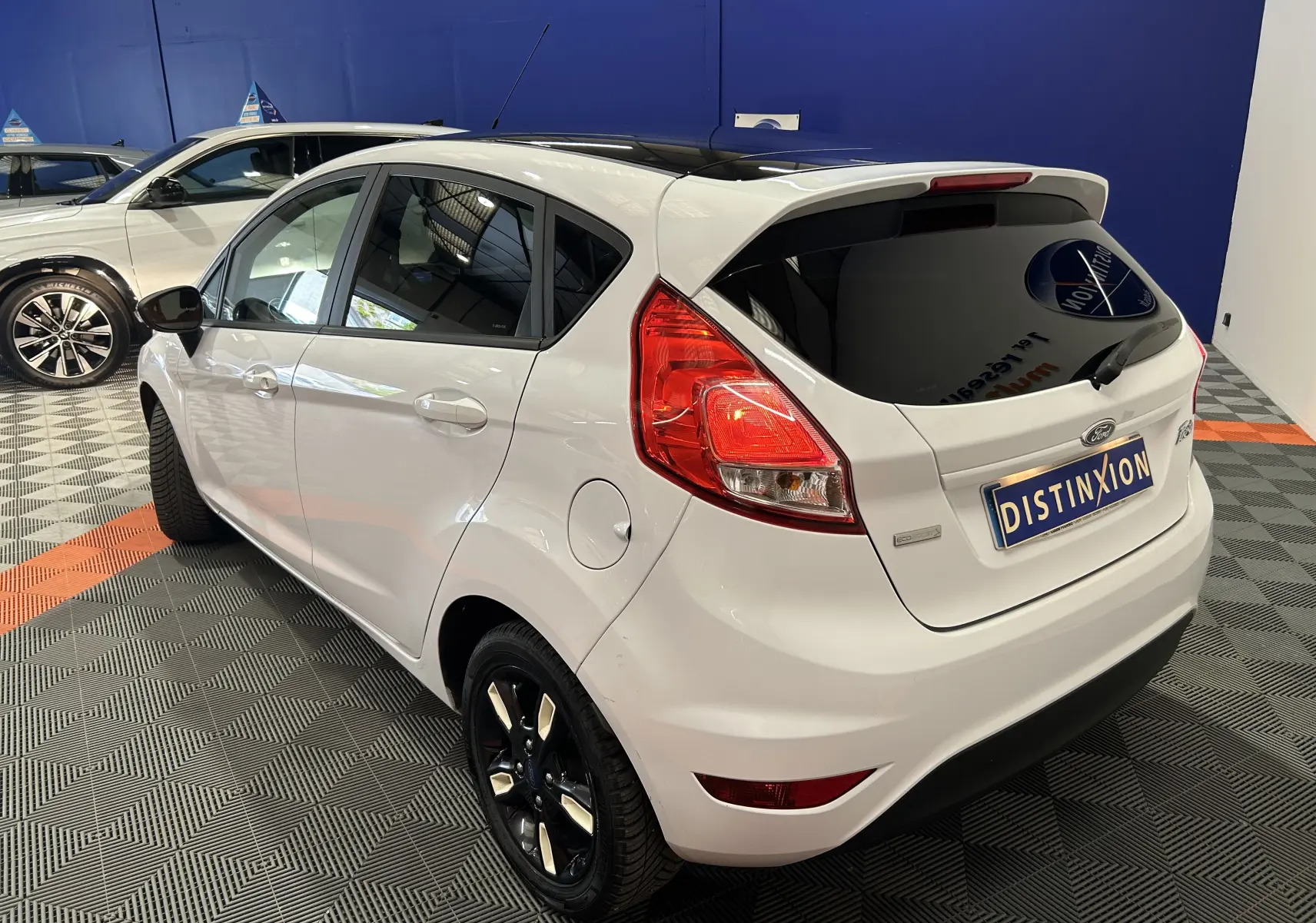 Vue 3/4 arrière droite d'une Ford Fiesta blanche avec jantes noires et vitres arrière teintées en intérieur showroom.