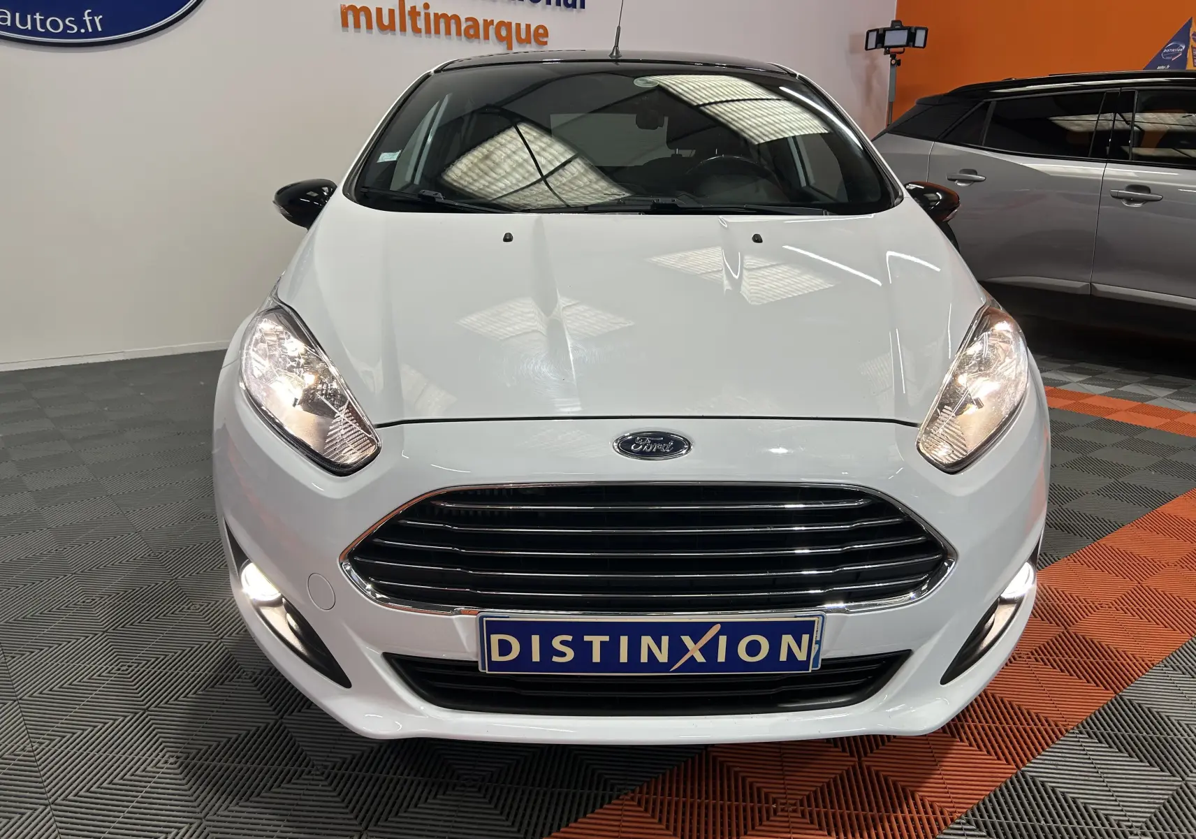 Vue avant d'une Ford Fiesta blanche EcoBoost 2015 avec phares allumés et calandre chromée dans un showroom.