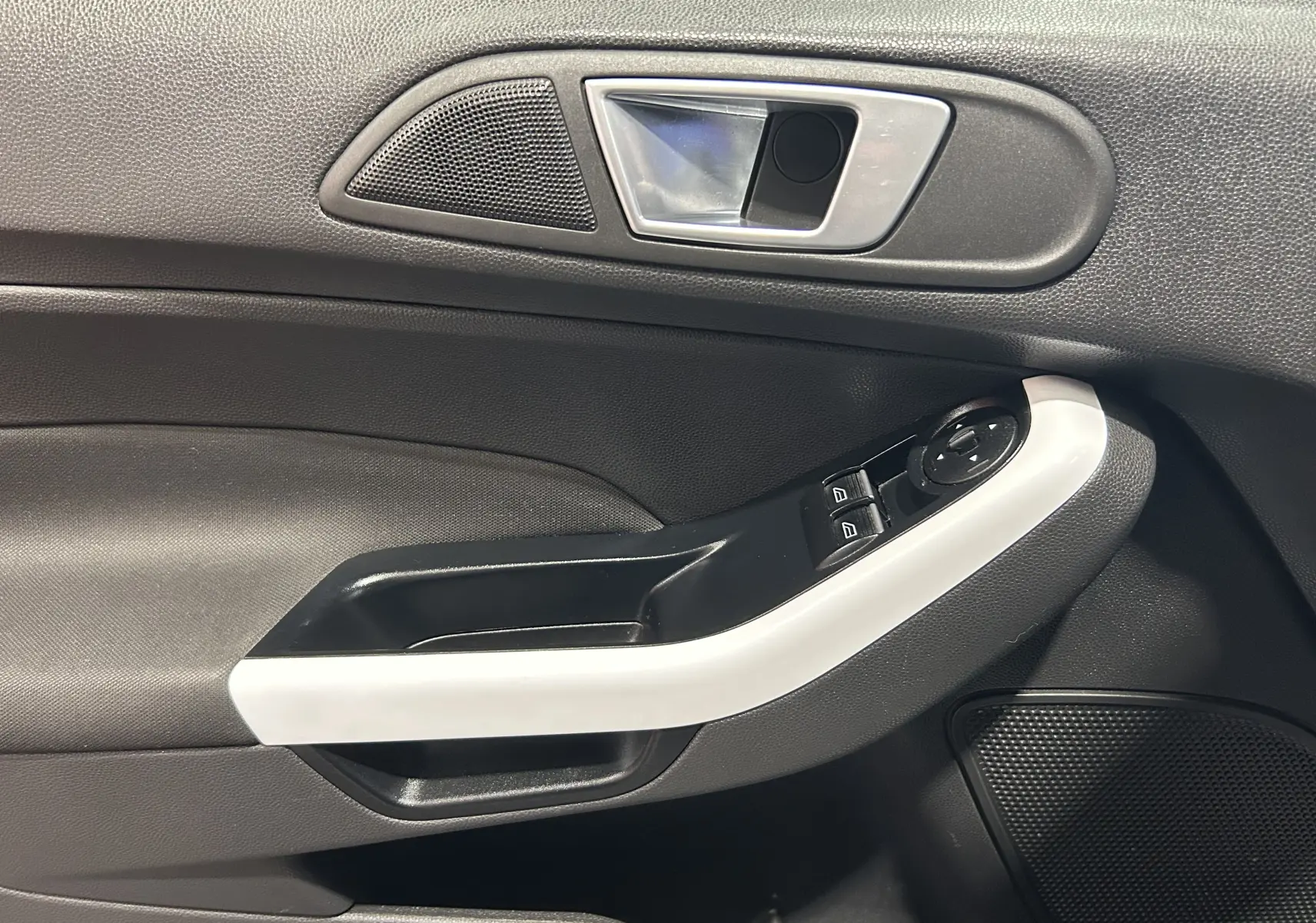 Gros plan sur la poignée intérieure côté gauche de la porte d’une Ford Fiesta blanche, avec commandes de vitres et haut-parleur intégré.