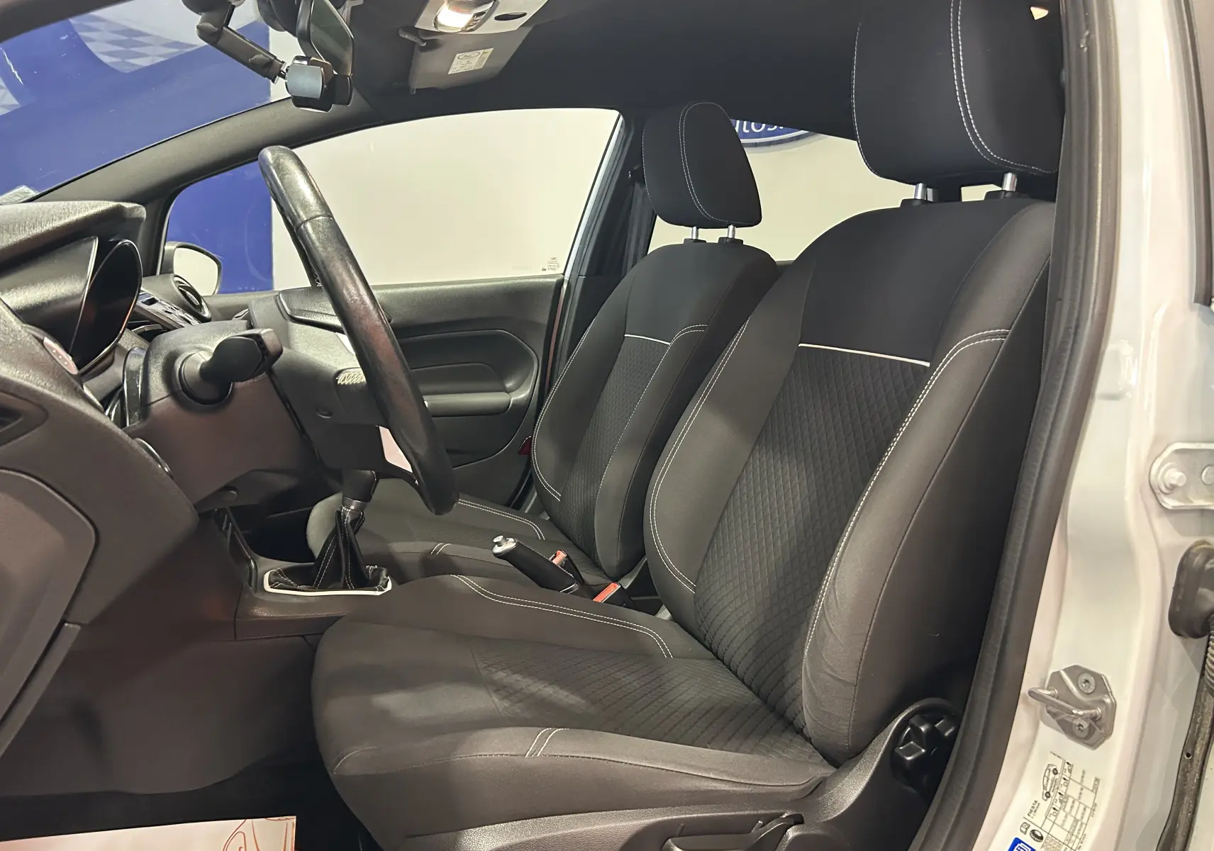 Vue intérieure côté gauche de la Ford Fiesta blanche 2015, montrant les sièges avant en tissu noir et le volant.