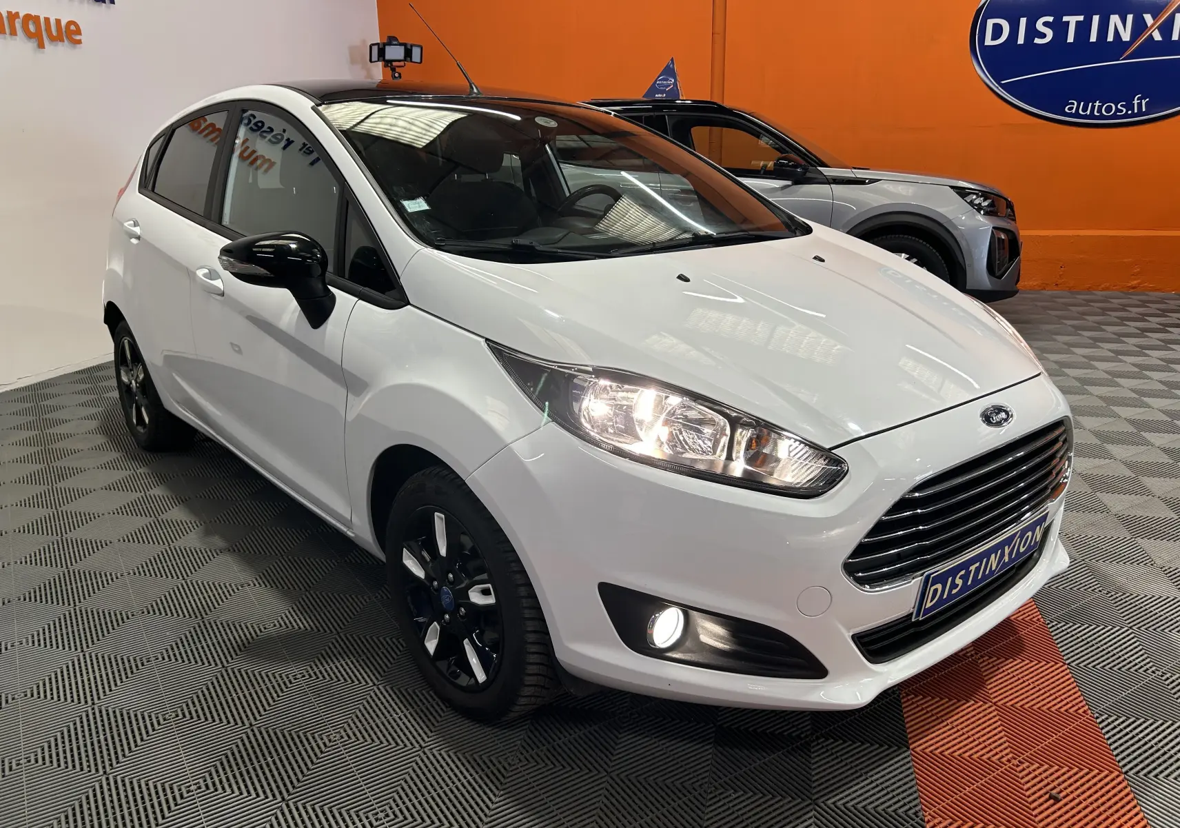 Vue 3/4 avant droite d'une Ford Fiesta blanche avec jantes noires et phares allumés dans un showroom.