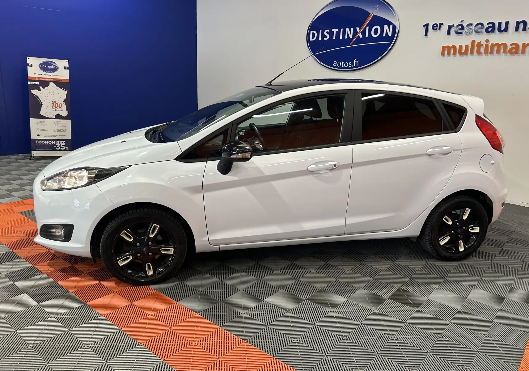 Vue de profil côté gauche d'une Ford Fiesta blanche avec jantes noires, stationnée en intérieur sur sol gris et orange.