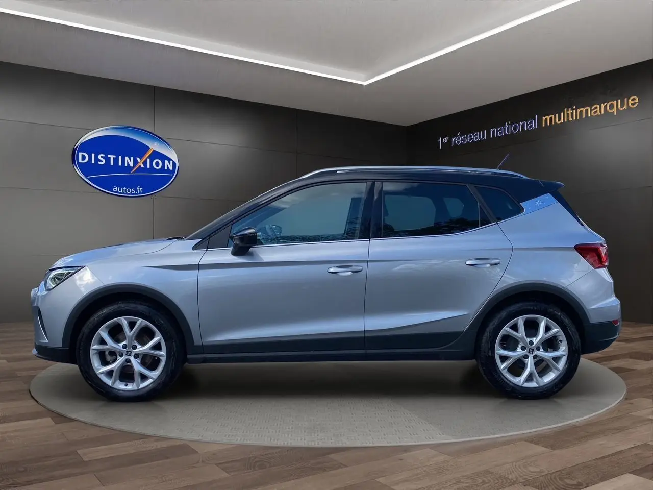 Vue de profil côté gauche d'un SEAT Arona gris silver avec toit noir et jantes alu 17 pouces dans un showroom.