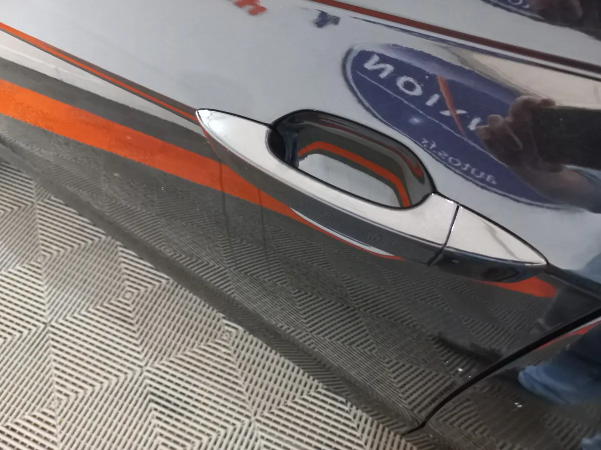 Poignée de porte côté gauche d'une Volkswagen Polo noire 2020 avec reflet d'un logo et sol à motifs géométriques.