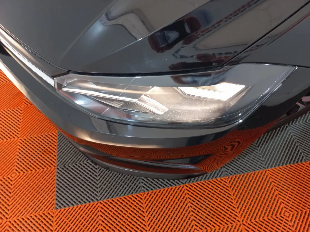 Gros plan sur le phare avant droit d'une Volkswagen Polo noire, avec reflets sur la carrosserie et sol orange et gris.