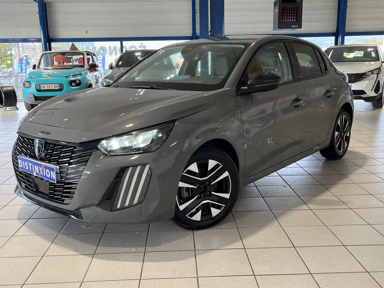 Peugeot 208 gris clair sélénium vue 3/4 avant avec calandre noire et jantes alliage bi-tons en showroom.