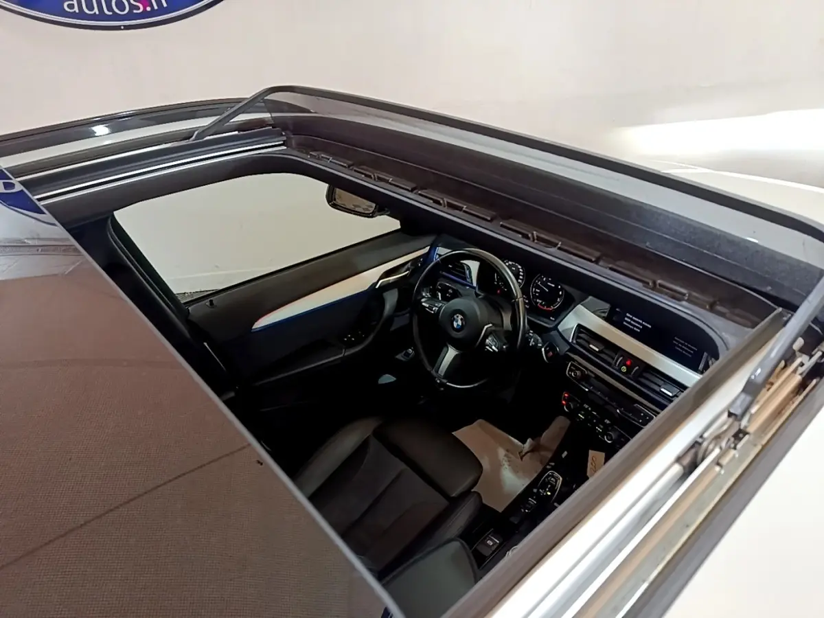 Vue intérieure par le toit ouvrant d'une BMW X1 blanc, montrant le tableau de bord et volant cuir multifonctions.