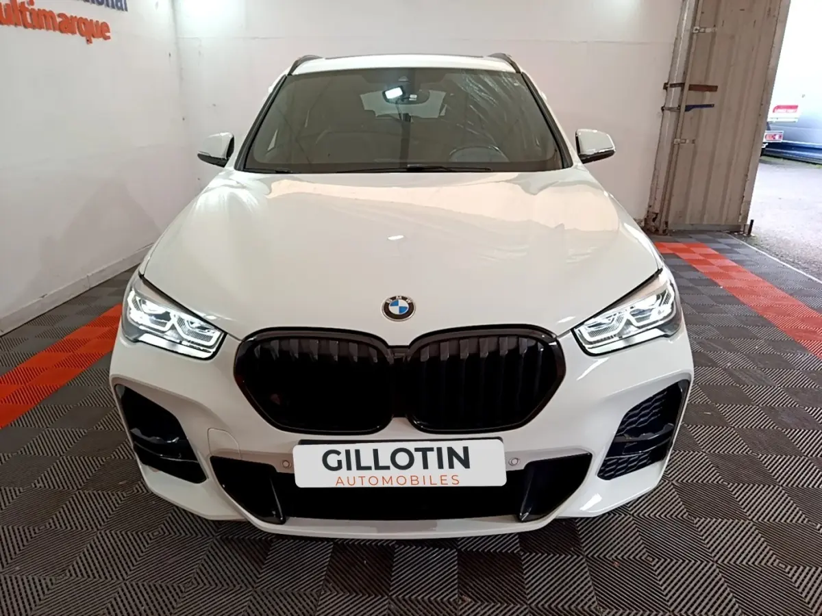 Vue avant d'une BMW X1 sDrive18i M Sport blanche avec calandre noire et phares LED allumés dans un showroom.