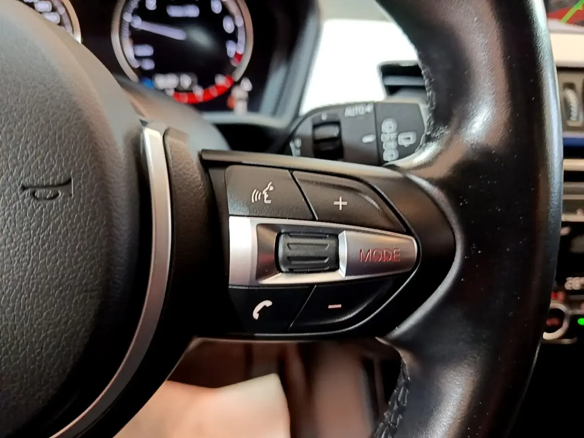 Gros plan sur la partie droite du volant noir en cuir du BMW X1 blanc, avec commandes audio et mode visibles.