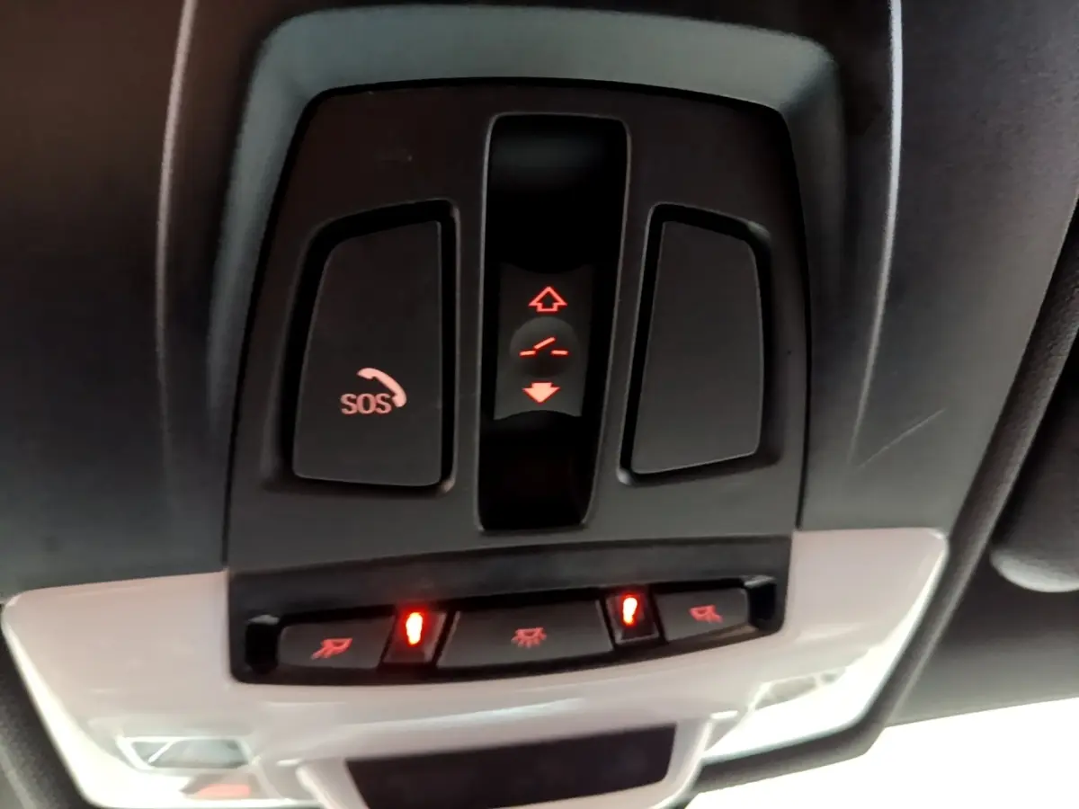 Vue en gros plan du panneau de commandes au plafond du BMW X1 blanc, avec bouton SOS et éclairage intérieur activé.