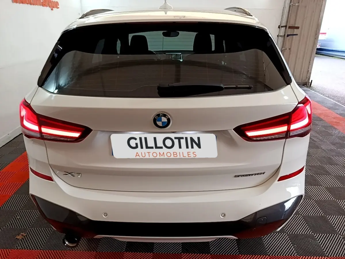 Vue arrière d’un BMW X1 blanc avec feux LED allumés, badge sDrive18i M Sport et double sortie d’échappement visible.