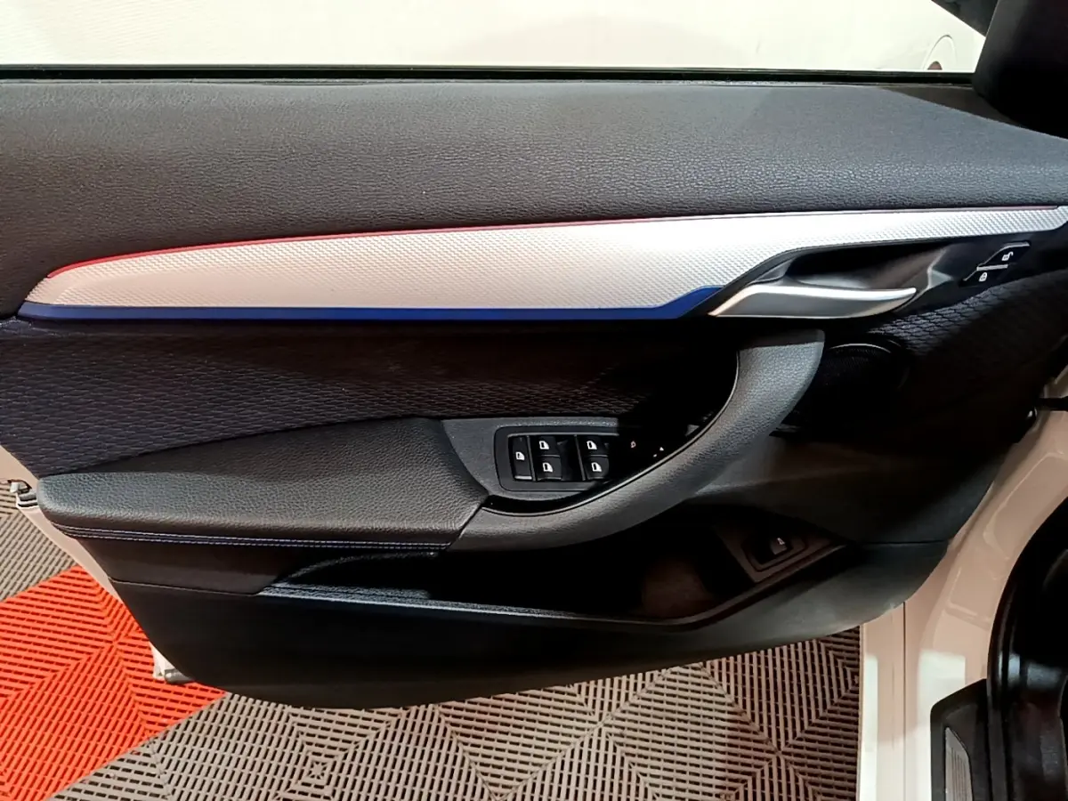 Vue intérieure côté gauche de la porte avant d’une BMW X1 blanche, avec insert décoratif tricolore et commandes de vitres.
