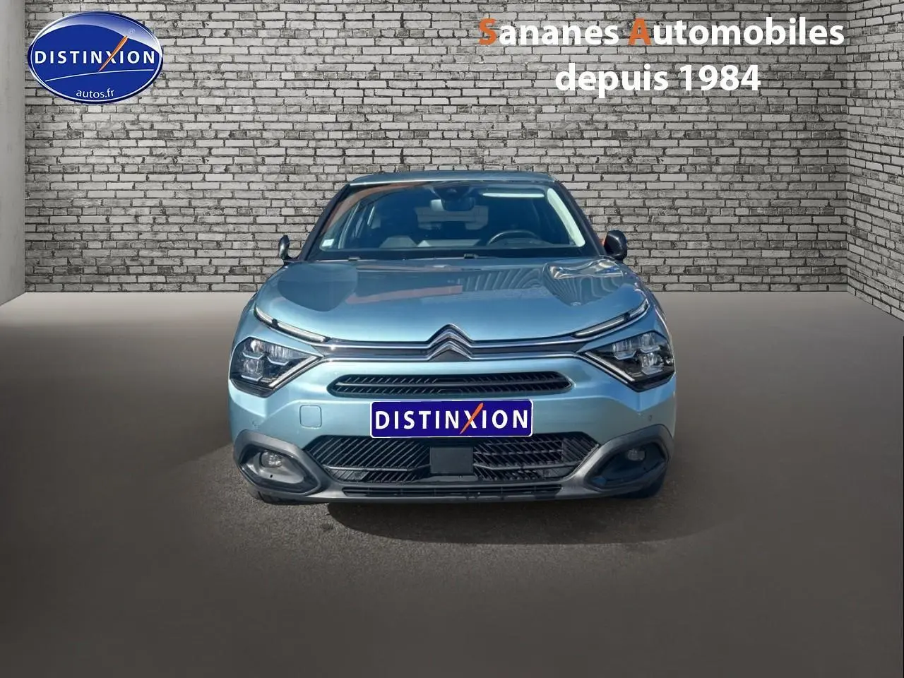 Vue de face d'une Citroën C4 bleu clair 2021 avec calandre distinctive et phares modernes, sur fond de mur en briques.