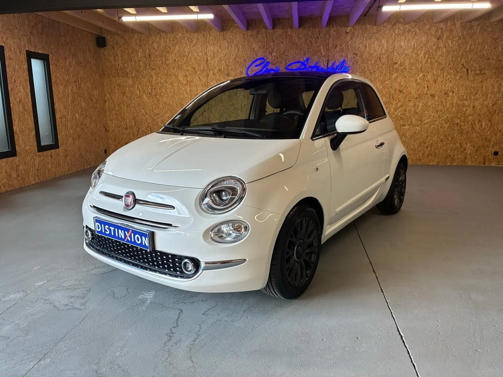 Fiat 500 blanc crème vue 3/4 avant droit dans un intérieur avec mur en bois et jantes noires.