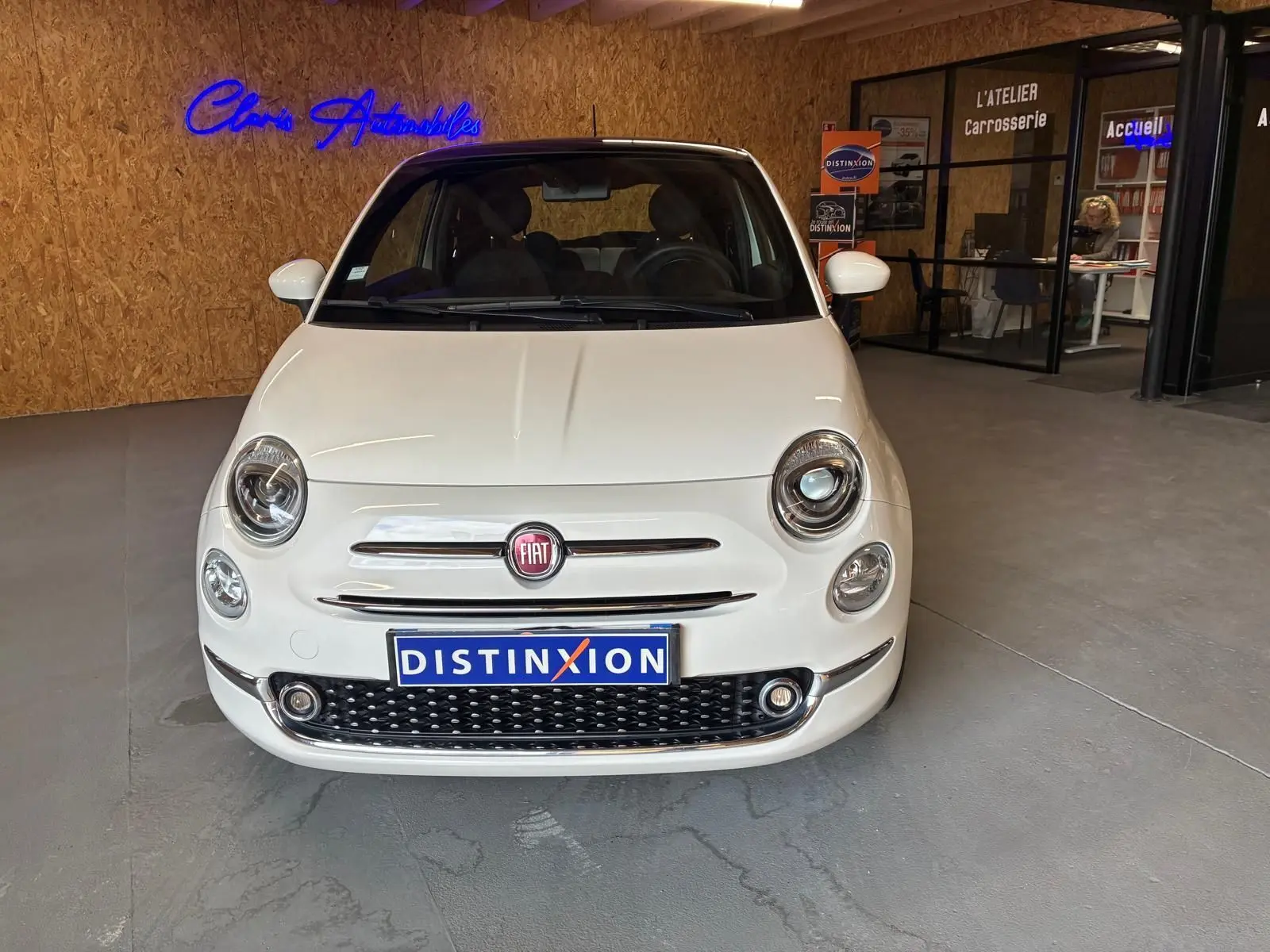 Vue de face d'une Fiat 500 blanc crème stationnée en intérieur, avec logo FIAT et plaque Distinxion visible.