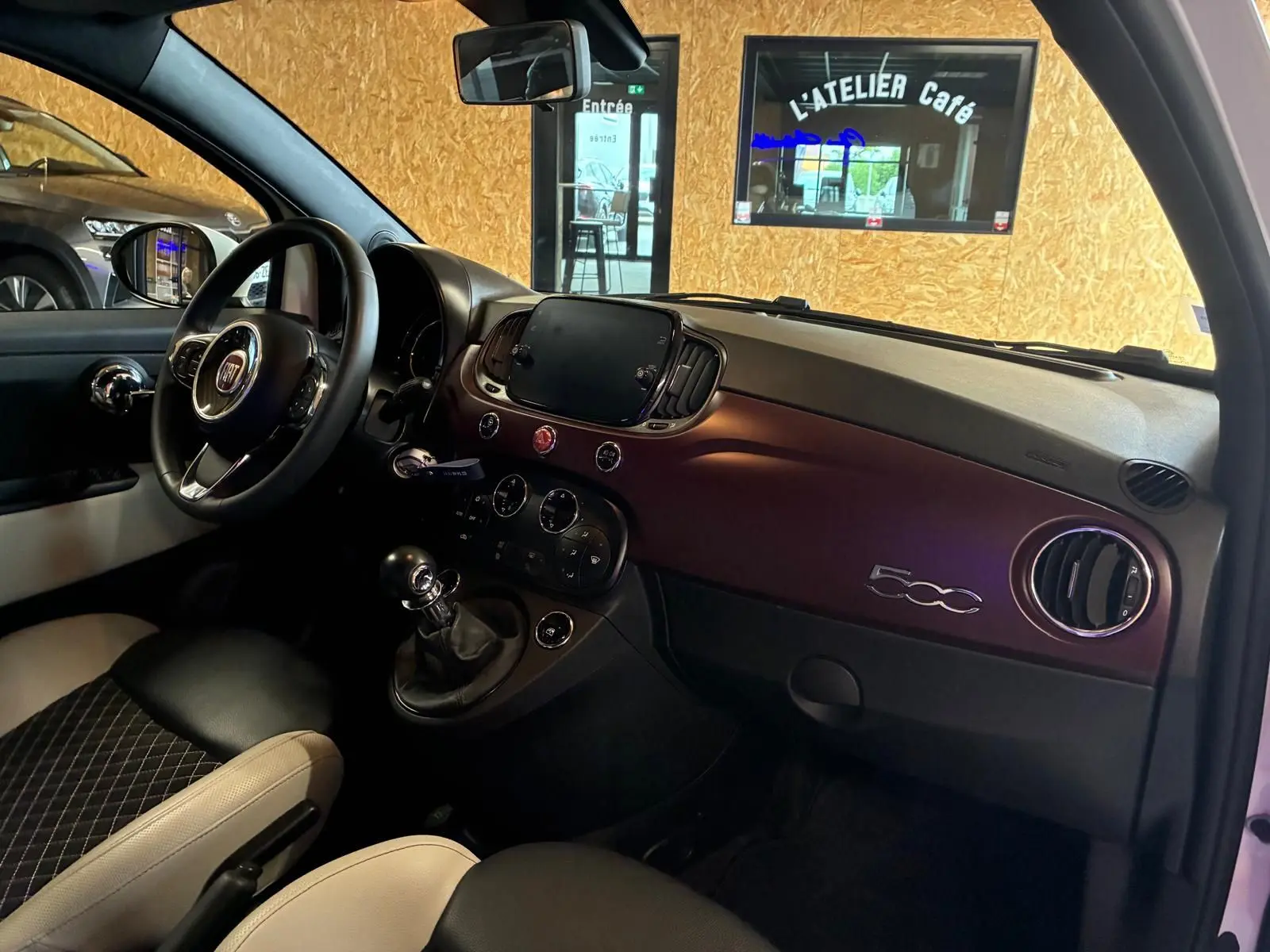 Vue intérieure côté conducteur de la Fiat 500 blanche crème, tableau de bord bordeaux avec logo 500 et volant noir.