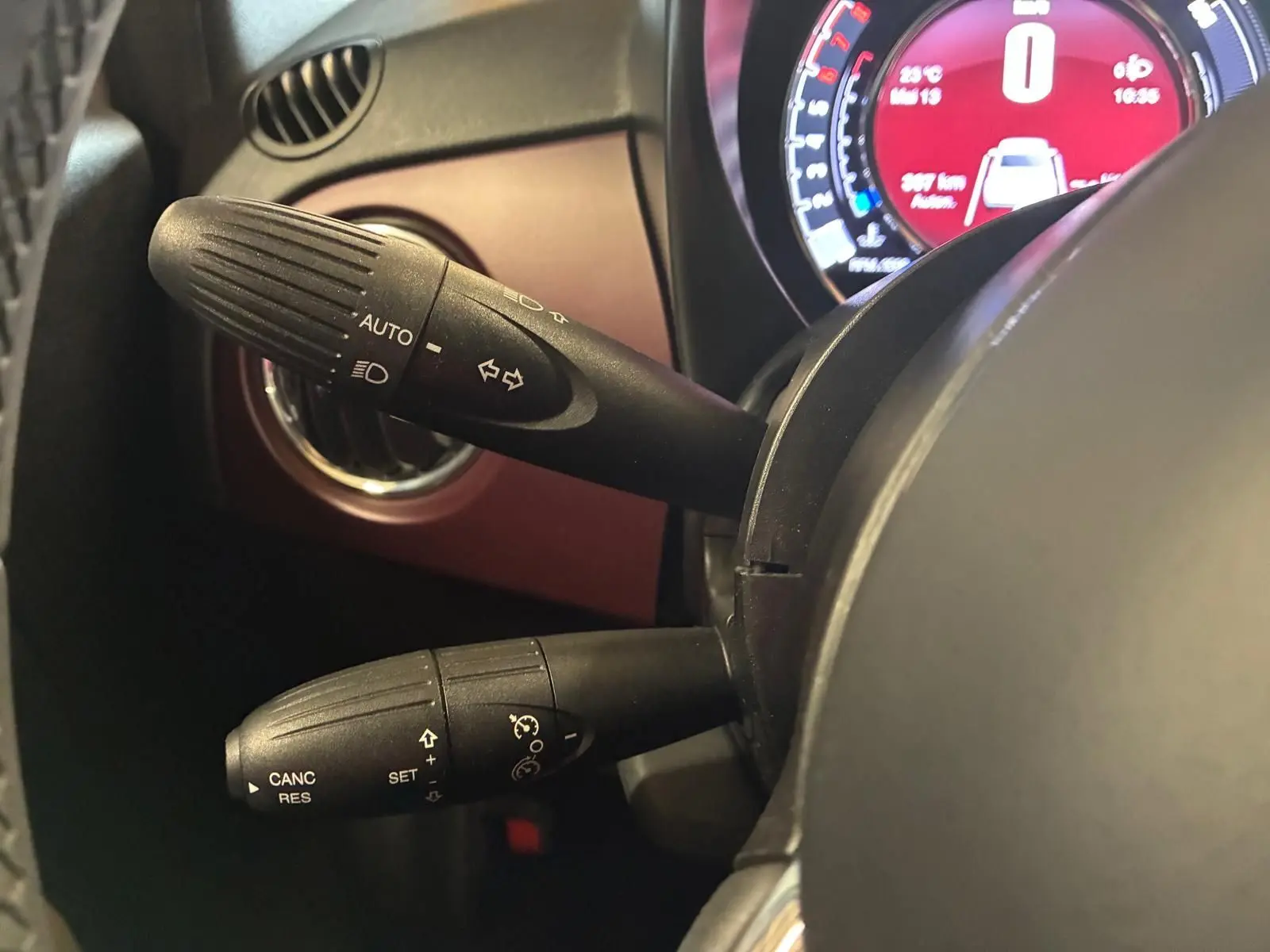 Gros plan sur les commandes de clignotants et régulateur de vitesse à gauche du volant d'une Fiat 500 blanc crème, tableau de bord numérique visible.