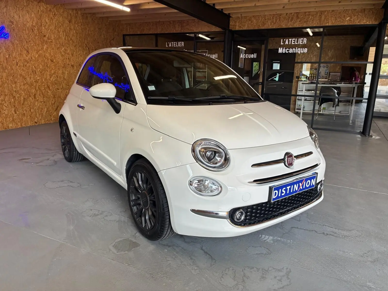 Vue 3/4 avant d'une FIAT 500 blanc crème, avec jantes noires et calandre chromée, stationnée en intérieur.