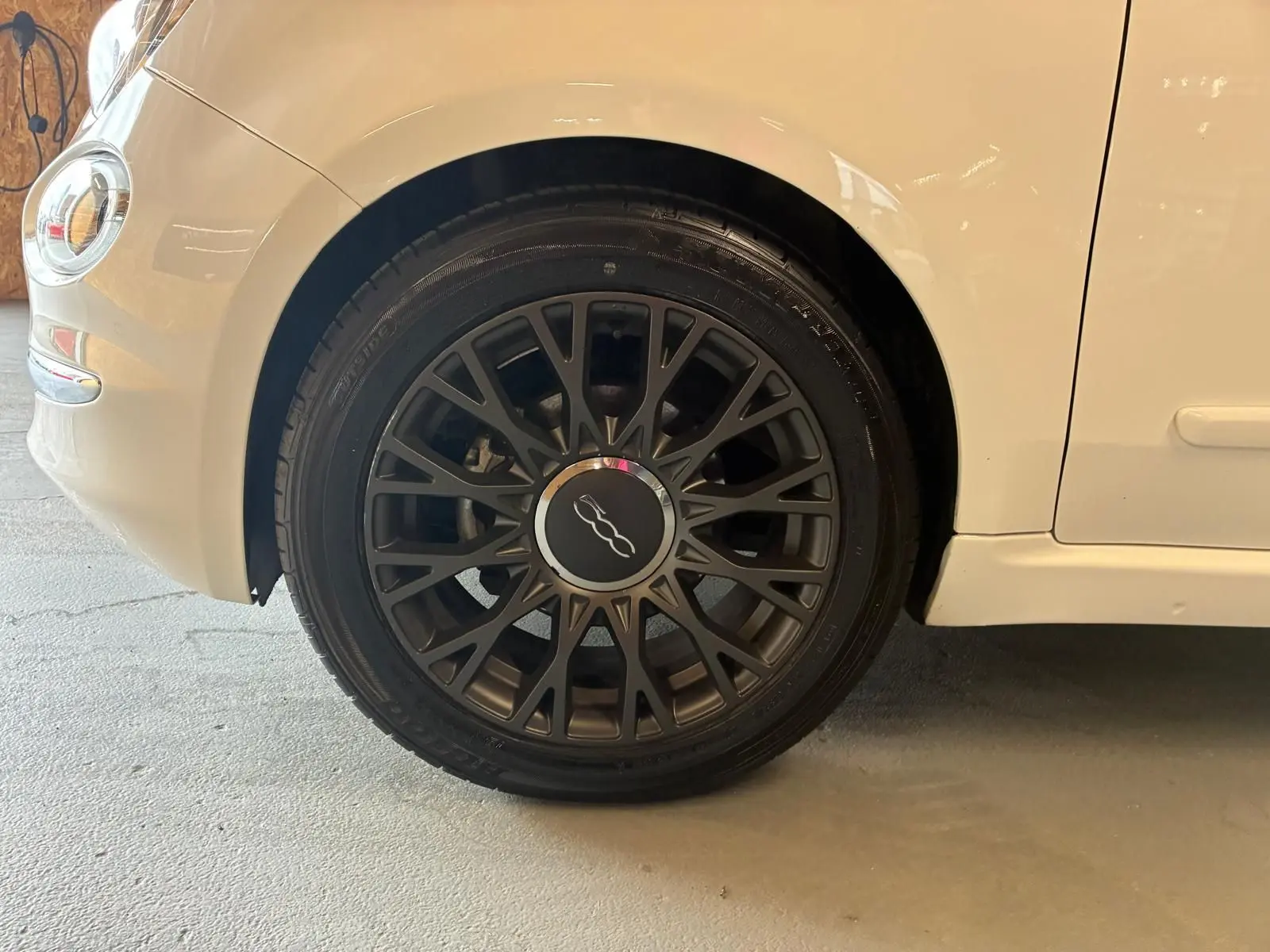 Gros plan sur la roue avant gauche noire à jante ajourée d'une FIAT 500 blanc crème vue de profil.