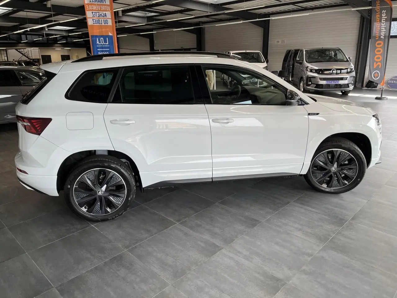 Profil côté gauche du SUV blanc SKODA KAROQ Sportline 2025 avec jantes alliage noires et toit noir en showroom.