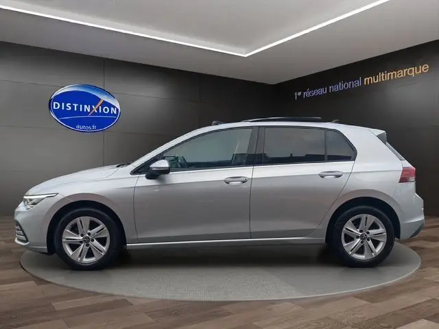 Profil côté gauche d'une Volkswagen Golf VIII gris argent avec vitres surteintées et toit panoramique ouvrant, en showroom.