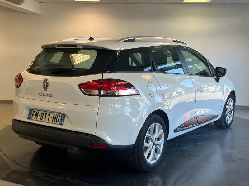 Vue 3/4 arrière droite d'une Renault Clio Estate blanche Glacier avec barres de toit et feux arrière rouges.