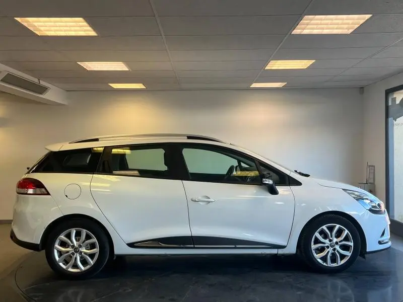 Profil latéral droit de la Renault Clio Estate blanc glacier avec barres de toit et jantes alliage en intérieur showroom.