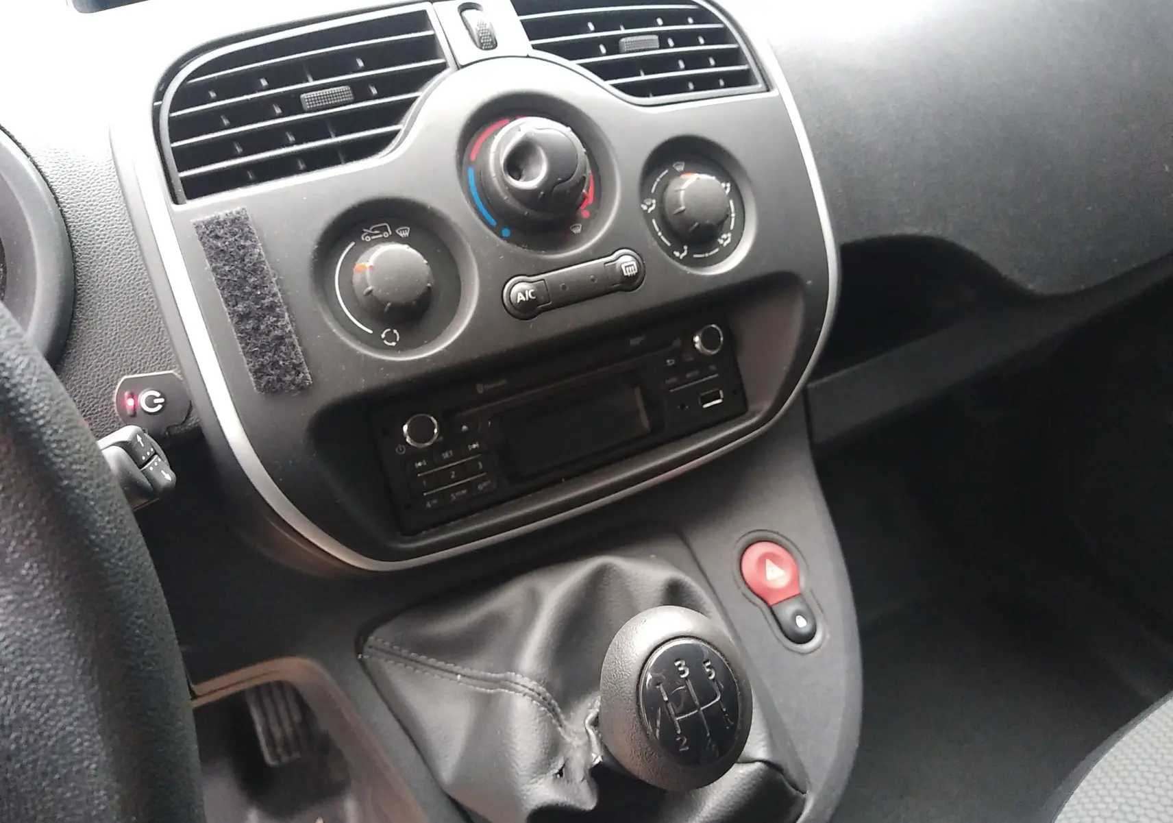 Vue rapprochée du tableau de bord et levier de vitesses manuel d'un Renault Kangoo blanc, avec commandes de climatisation et radio intégrées.