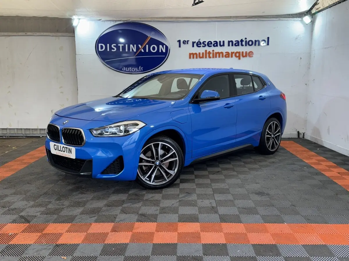 BMW X2 xDrive25e M Sport bleu en 3/4 avant droit dans un showroom avec sol gris et bandes orange.