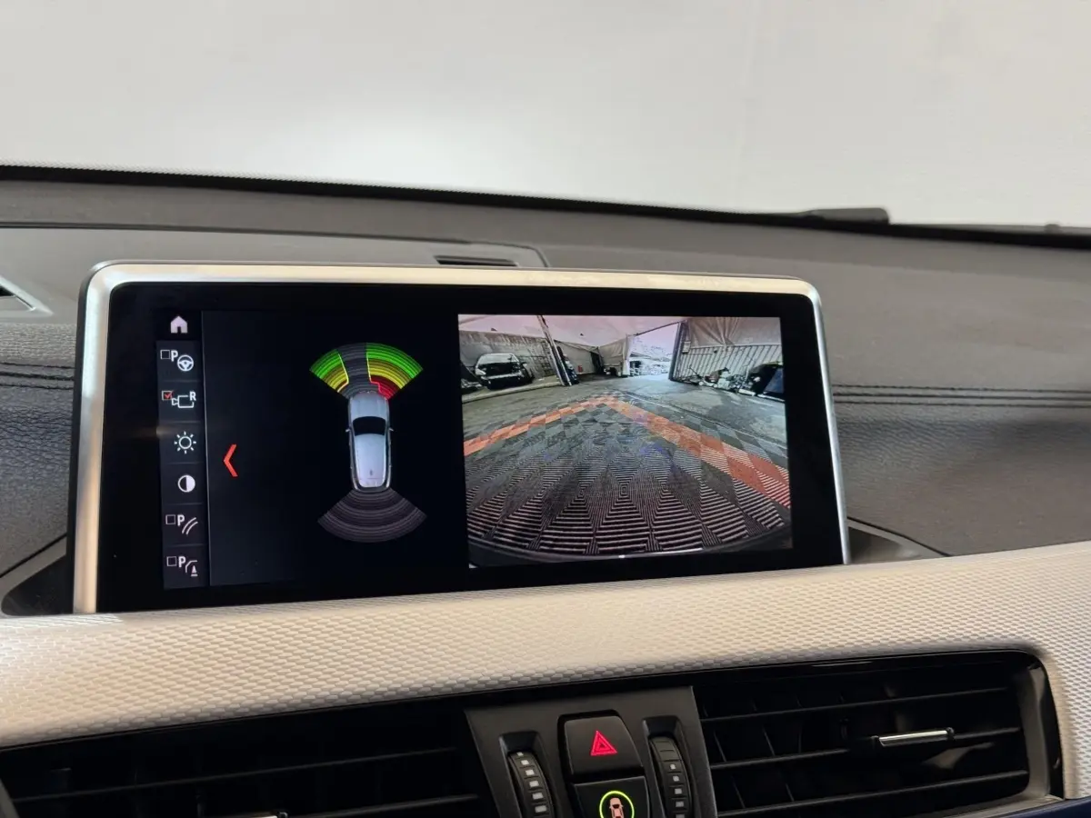 Écran central de BMW X2 xDrive25e bleu montrant l’aide au stationnement avec vue caméra arrière et capteurs.