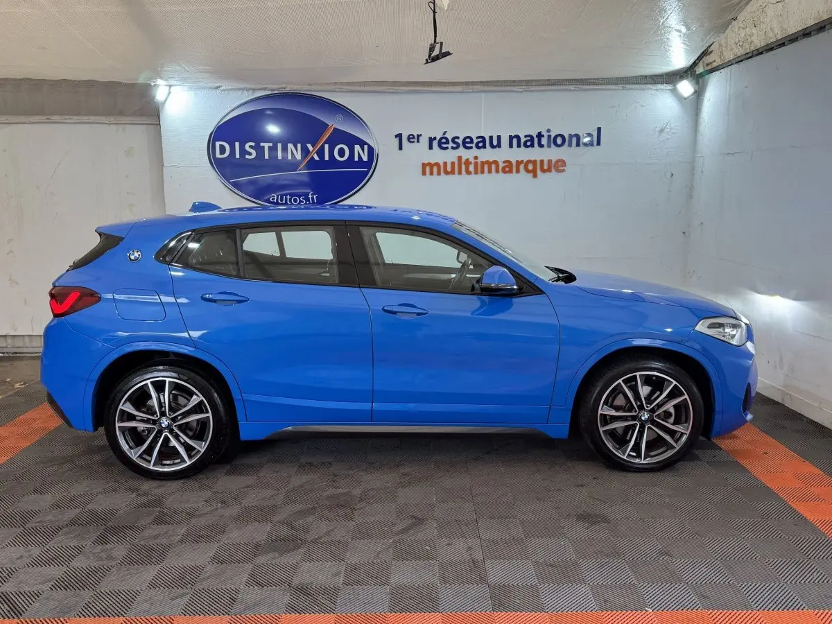 Vue de profil côté gauche d’un BMW X2 xDrive25e M Sport bleu dans un garage avec logo Distinxion en arrière-plan.