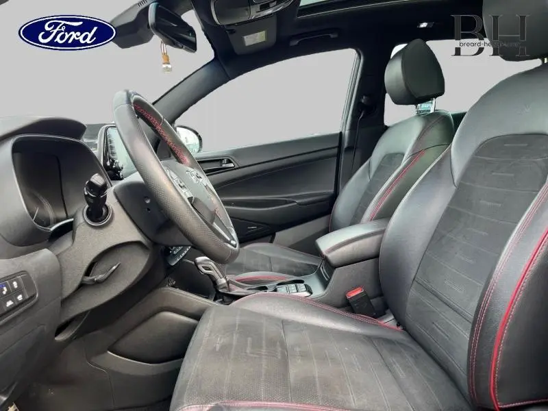 Vue intérieure côté conducteur d'un Hyundai Tucson 2020 avec sièges noirs en cuir et surpiqûres rouges, volant multifonction.