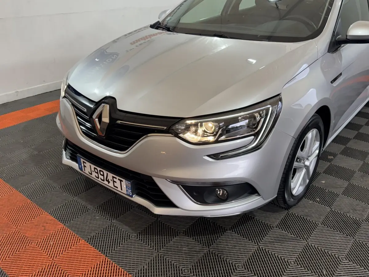 Renault Mégane Business gris clair vue 3/4 avant droit, phare allumé et calandre noire distinctive.