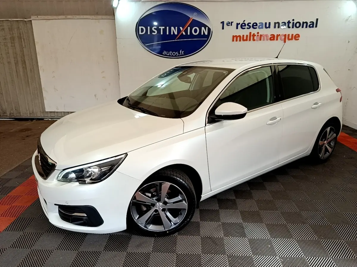 Peugeot 308 blanc vue 3/4 avant droit en intérieur, avec jantes alliage et phares ellipsoïdaux visibles.