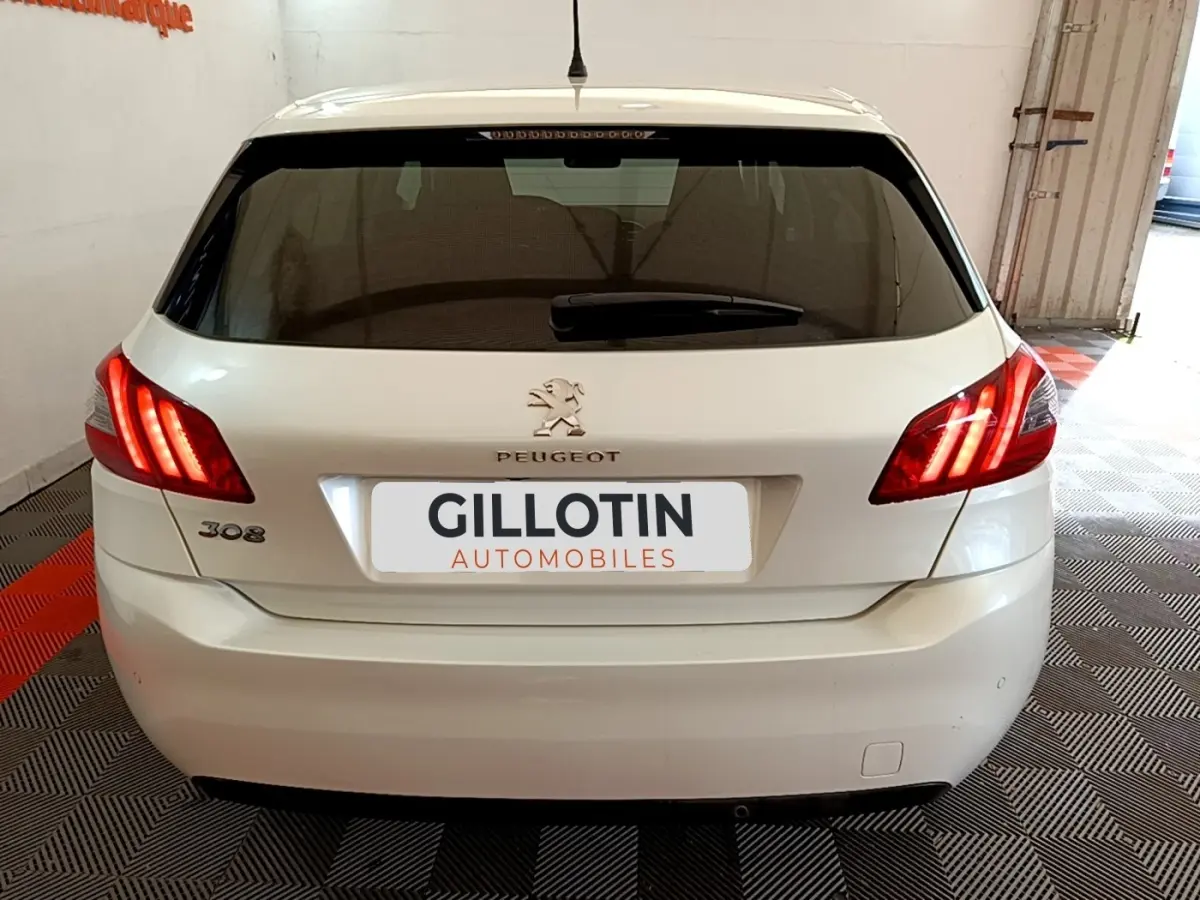 Vue arrière d'une Peugeot 308 blanche de 2017 avec feux arrière allumés et logo visible sur un sol carrelé.