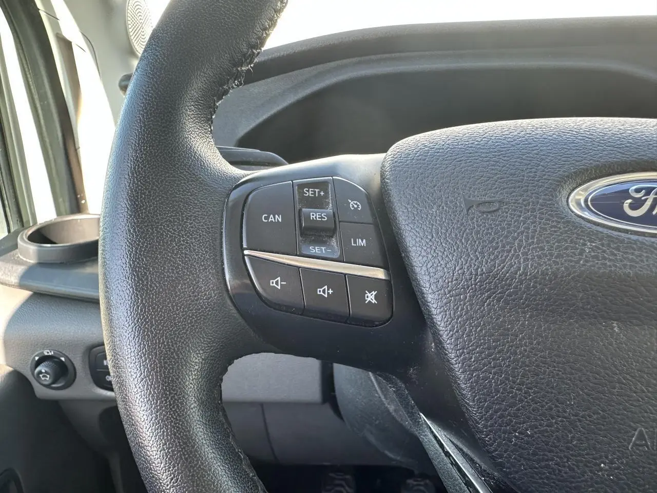 Gros plan sur le volant en cuir noir du Ford Transit Fourgon blanc, avec commandes de régulateur et volume audio.