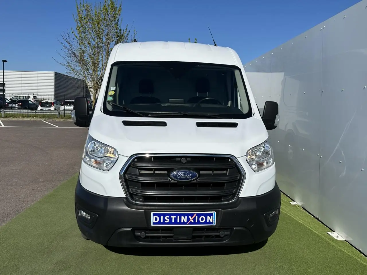 Vue frontale d'un fourgon Ford Transit blanc avec calandre noire et plaque Distinxion sur un parking extérieur.