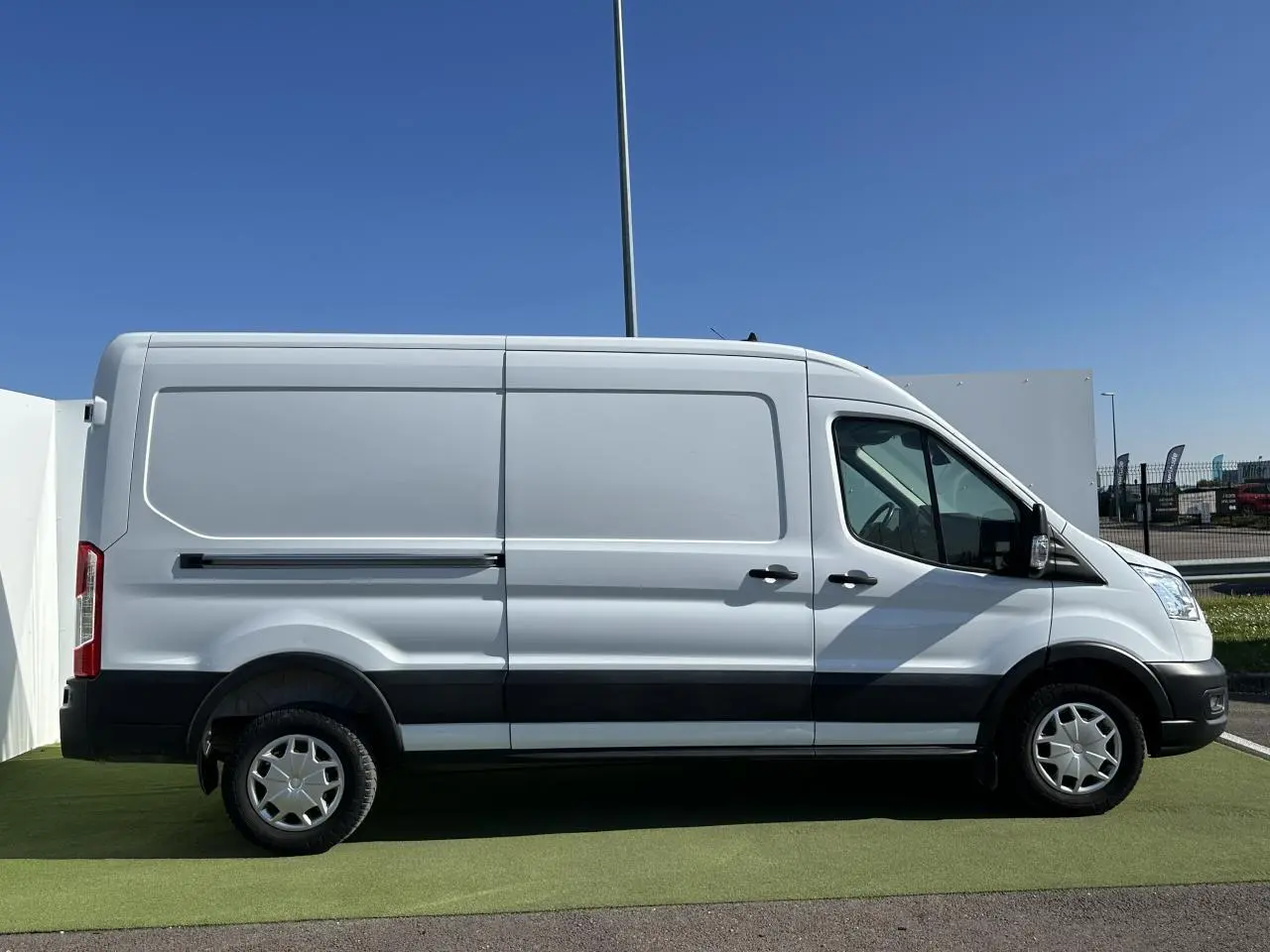 Ford Transit Fourgon blanc vu de profil côté gauche, avec bandes noires sur les bas de caisse et enjoliveurs pleins.