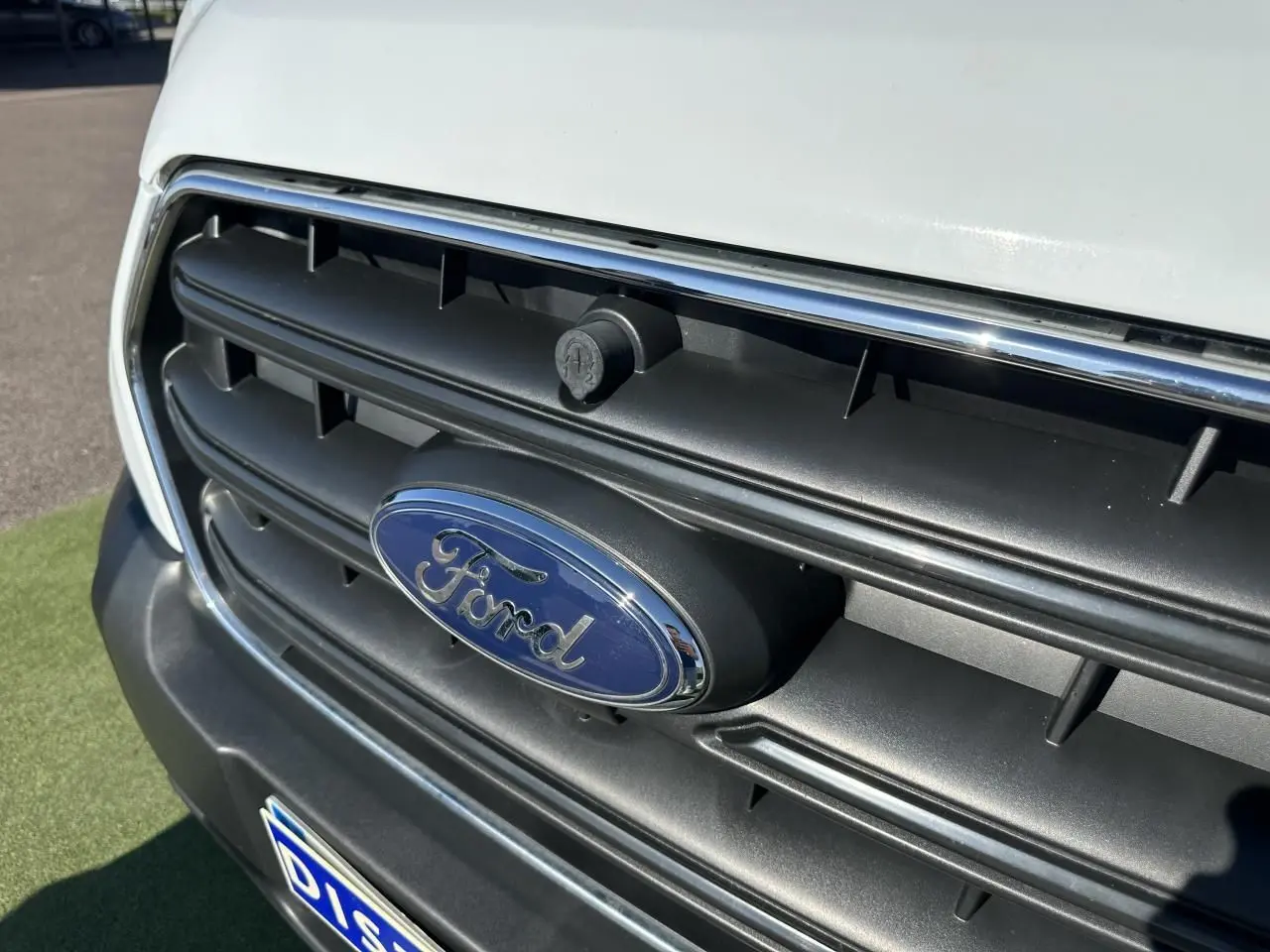 Gros plan sur la calandre noire avec logo Ford bleu et carrosserie blanche du Ford Transit Fourgon 2020.