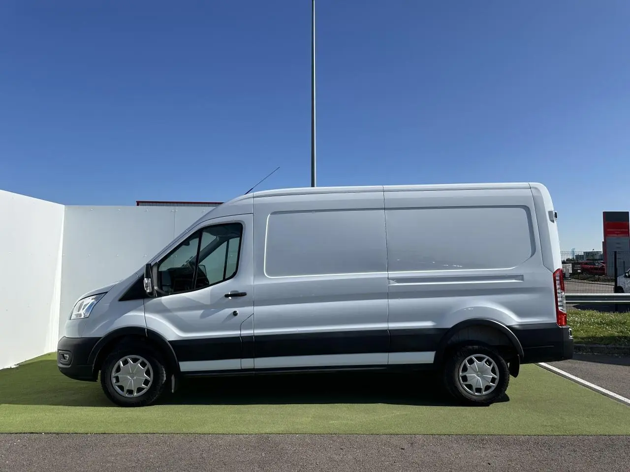 Profil côté gauche d'un fourgon Ford Transit blanc avec jantes acier et bande noire sur bas de caisse.