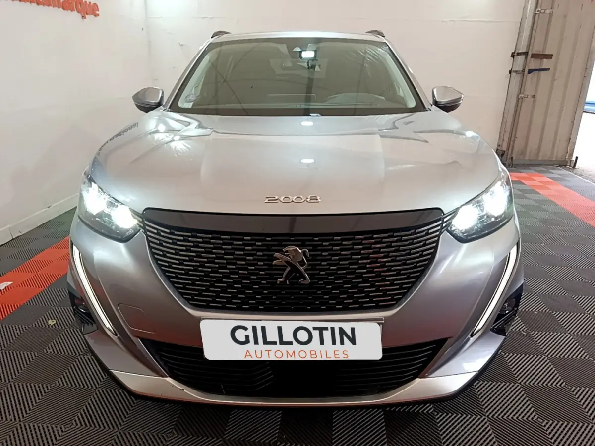 Vue frontale d'un Peugeot 2008 gris clair avec phares allumés et calandre noire distinctive en intérieur showroom.