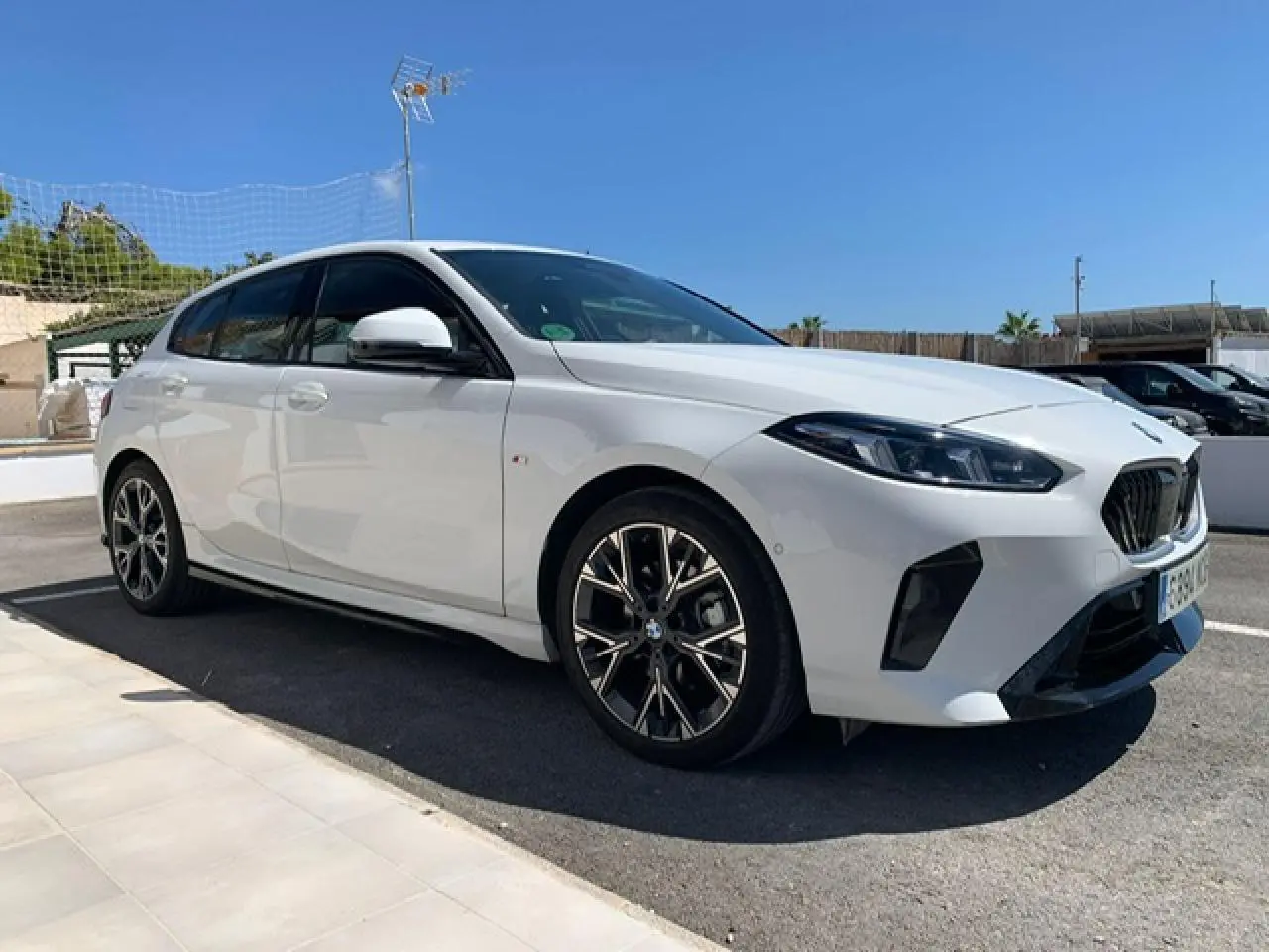 BMW Série 1 blanche vue 3/4 avant droit, avec jantes alliage bicolores et calandre M Sport sous un ciel bleu.